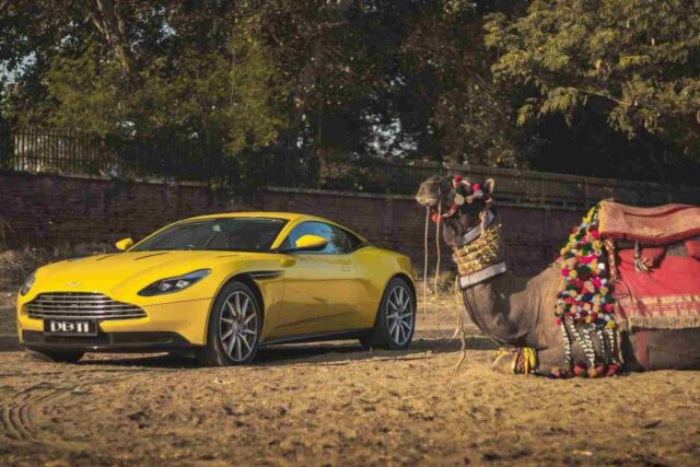 Çöl ortamında bir Aston Martin ve bir deve yan yana.
