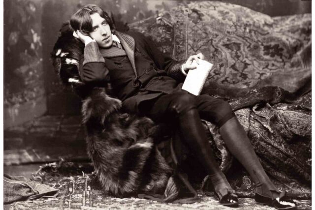 Oscar Wilde oturmuş, elinde bir defterle düşünüyor.